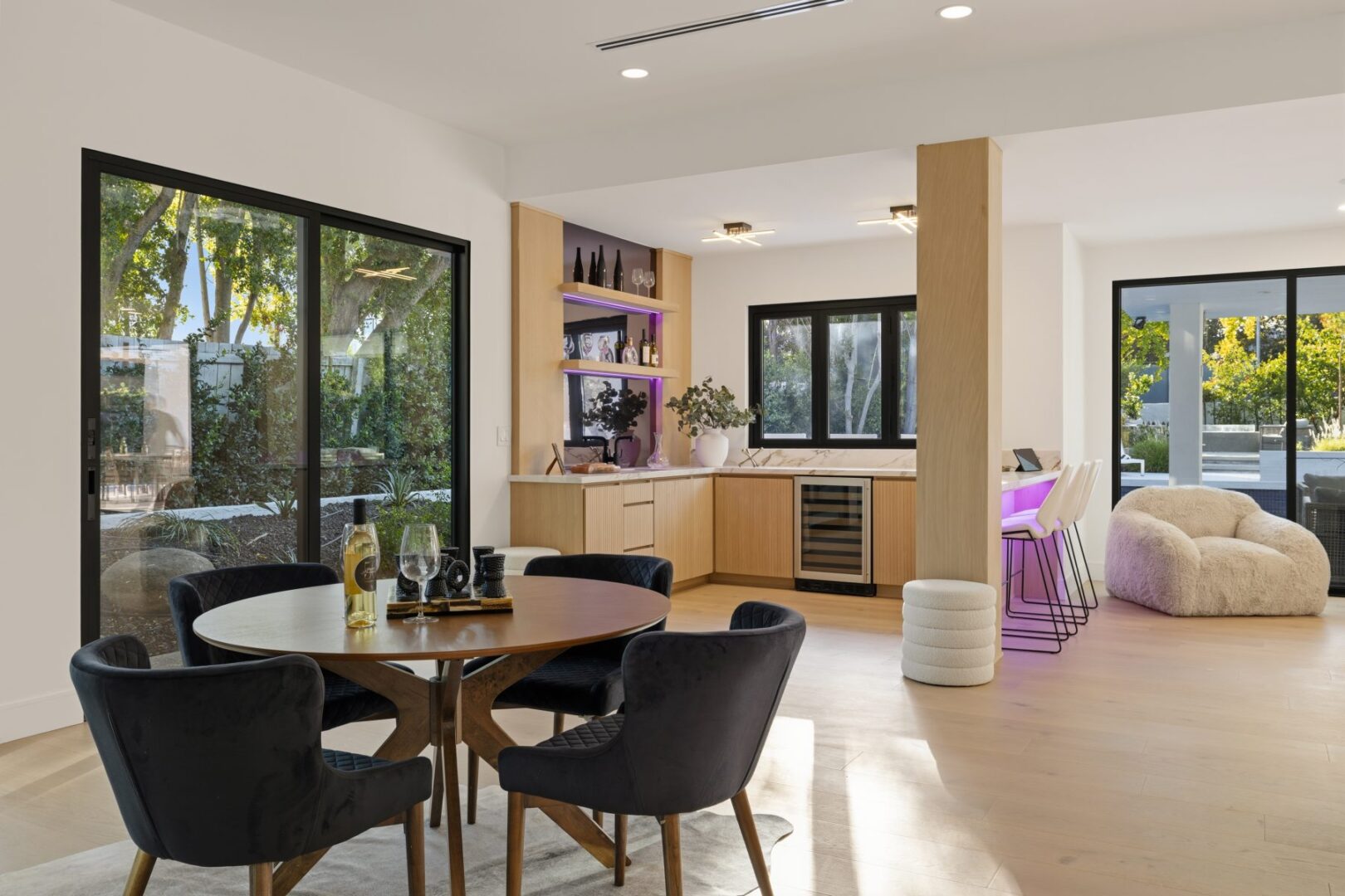 Dining table and dark chairs sit beside the kitchen, with sliding doors opening to a landscaped yard.