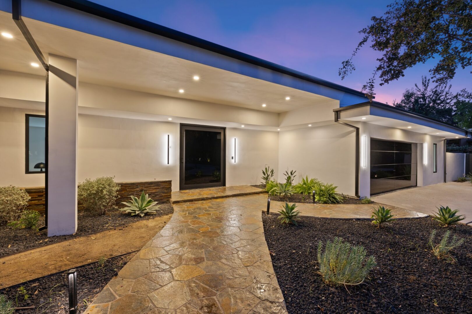 Covered front entry with a dark door, stone path, and landscaped beds beside the garage.