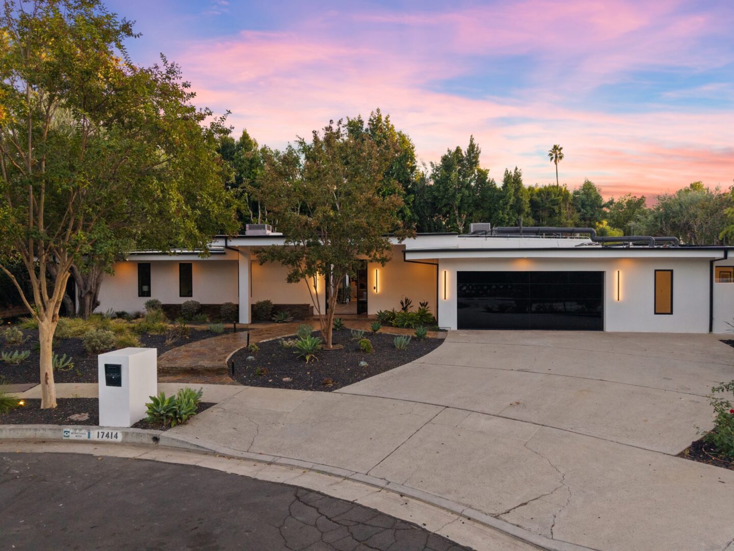 Low-profile home with a black garage door, front walkway, planted beds, and an entry set beneath trees.