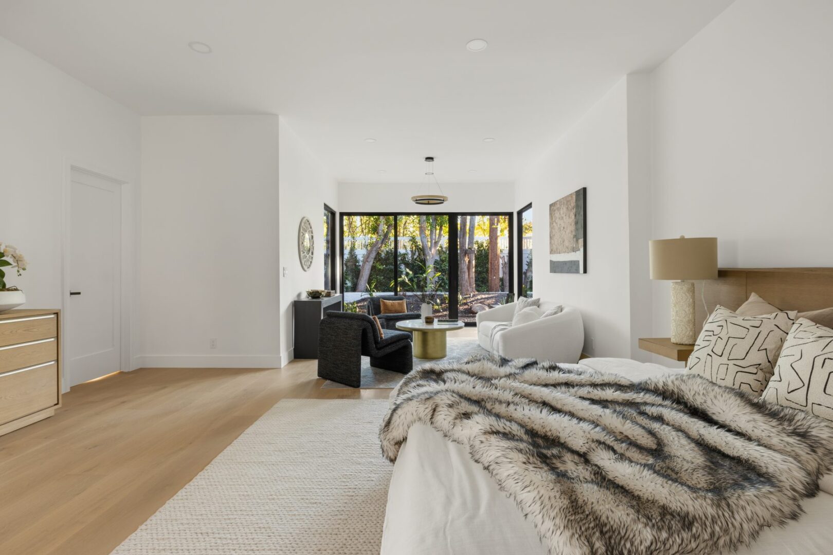 Living area with a bed in the foreground, seating by black-framed windows, and a garden view.