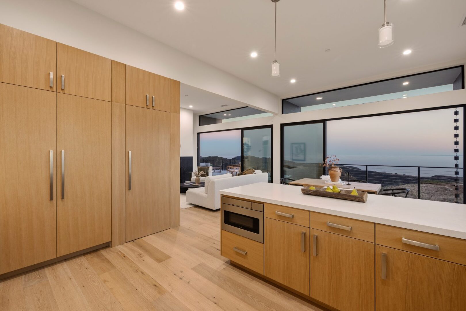 Modern kitchen with wooden cabinets, white countertops, and ocean view through large windows.