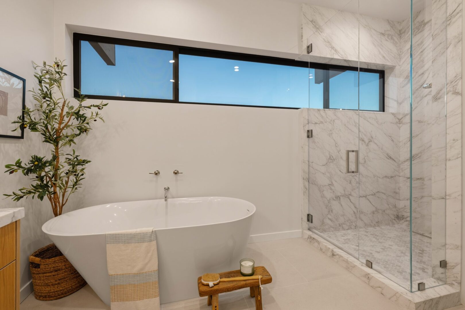 Bright bathroom with a freestanding tub and glass shower, featuring marble walls and a plant.