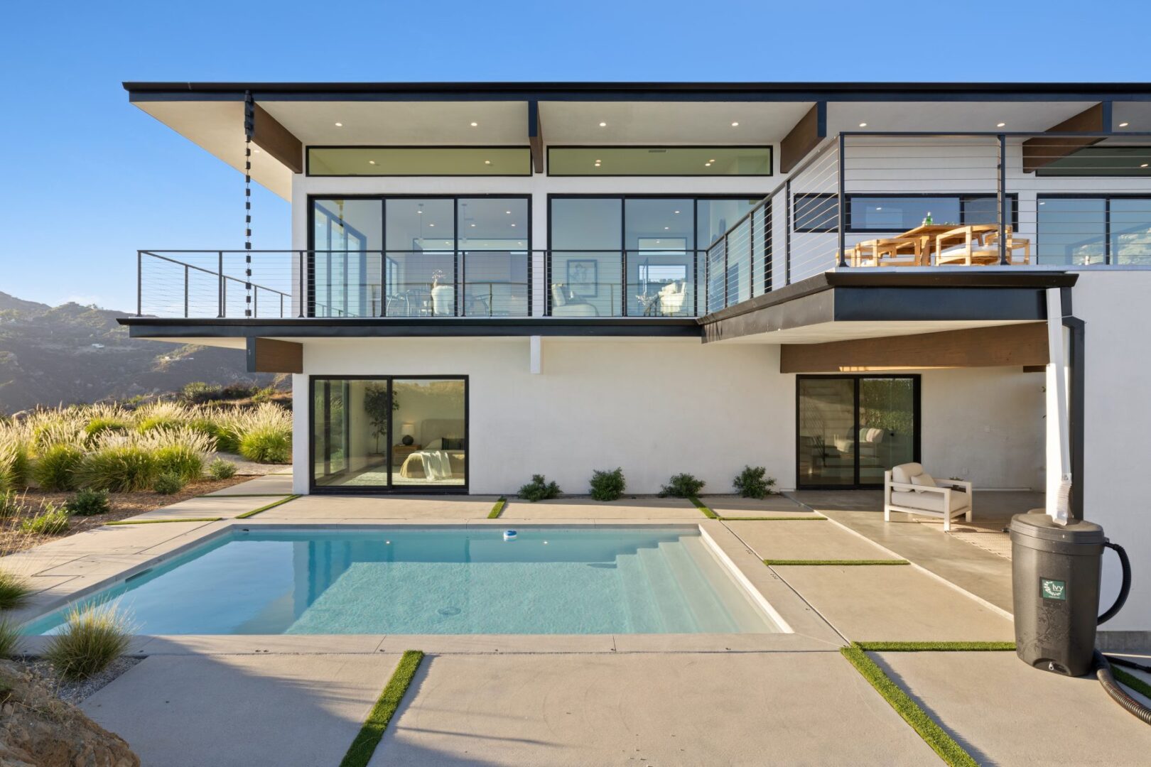 Modern home with pool and large windows, surrounded by landscaping under clear blue sky.