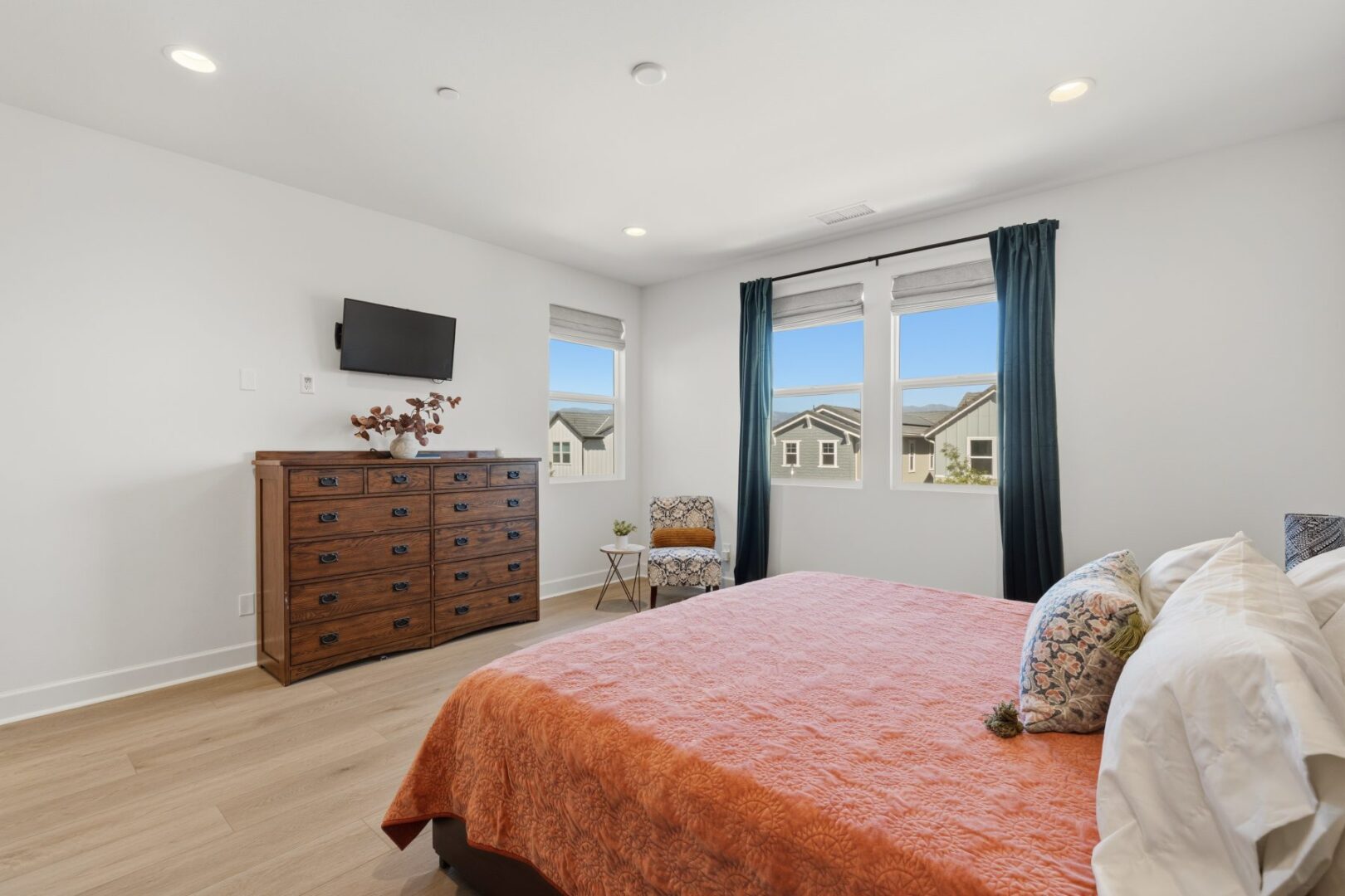 Bedroom with a dresser, wall-mounted television, upholstered bed, and windows overlooking neighboring homes.