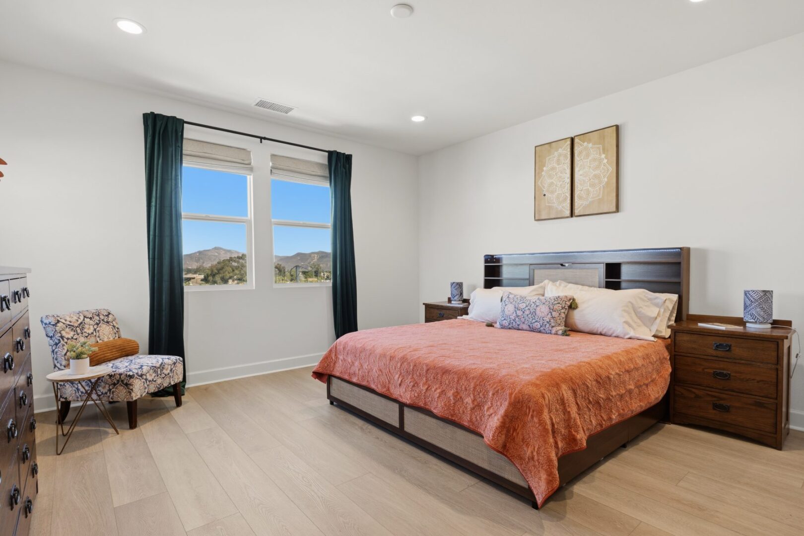 Bedroom with a large bed, wood nightstands, a patterned chair, and windows overlooking hills.